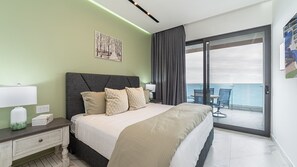 Une chambre d’hôtel moderne avec un grand lit, une table de chevet, une lampe, un tableau et une vue sur la mer, visible à travers une porte-fenêtre coulissante.