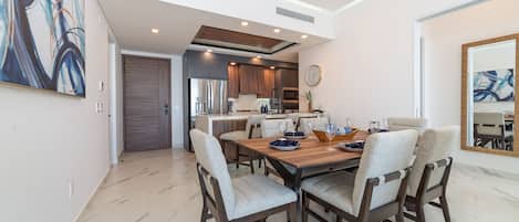A modern dining area with a wooden table, upholstered chairs, and a kitchen in the background.