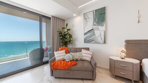 Ein modernes Wohnzimmer mit Balkon und Meerblick, ein graues Sofa mit Zierkissen, ein Beistelltisch mit Lampe sowie ein großes abstraktes Gemälde an der Wand.