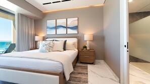 Quarto de hotel moderno com uma cama grande, mesa de cabeceira, luminária e vista para o oceano.