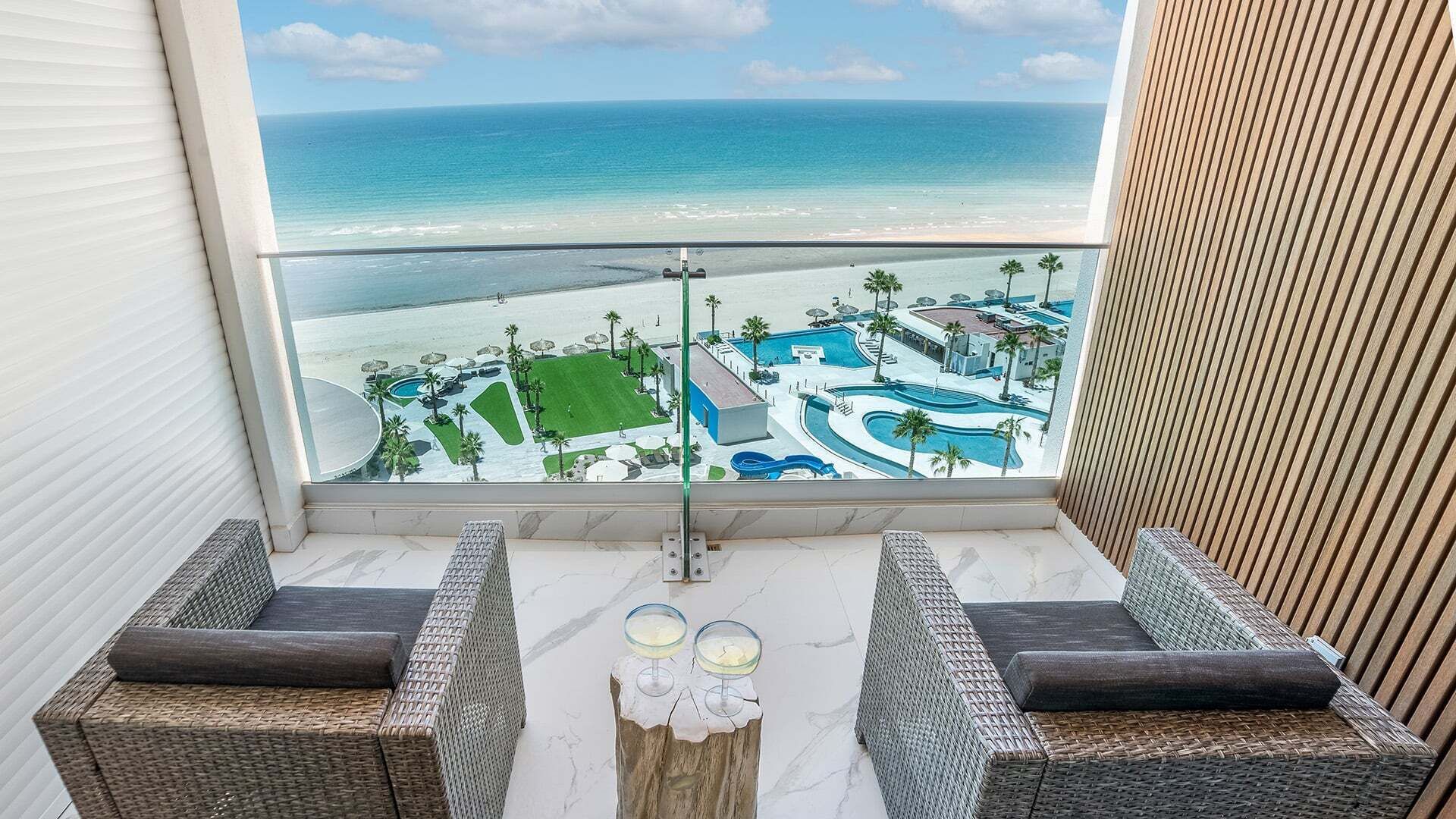 A balcony with wicker furniture, a view of a resort with a pool and beach, and a clear sky.