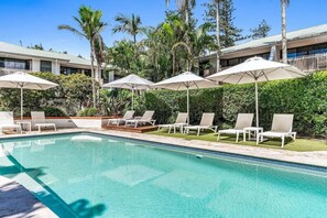 A swimming pool with lounge chairs and umbrellas, surrounded by greenery and a building with a white facade. - Sea Crest- Unit 5, 47 Shirley St, The Crest Apartments (Byron)