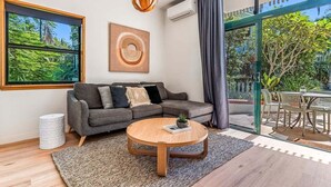 A modern living room with a grey sectional sofa, a round wooden coffee table, a large window with a view of greenery, and a ceiling light fixture. - Sea Crest- Unit 5, 47 Shirley St, The Crest Apartments (Byron)