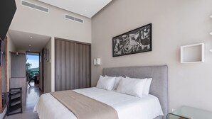 A modern bedroom with a large bed, a headboard, a bedside table, a framed artwork, and a view through an open door. - Luxury Oceanfront 2BR Maritima (Flamingos)