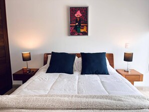 A neatly made bed with white bedding and two dark pillows, flanked by wooden nightstands with lamps, in a room with a single piece of wall art above the bed. - Vista al Caribe · 4 balcones · Plaza Marina · TOP (Cancún)