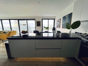 A modern kitchen with a large island, a dining area with yellow chairs, and a view of the cityscape through large windows. - Vista al Caribe · 4 balcones · Plaza Marina · TOP (Cancún)