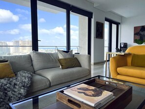 Uma sala de estar moderna com sofá, poltrona e mesa de centro, com uma grande janela e vista para a cidade.