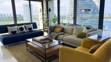 Un salon moderne avec un canapé, un fauteuil et une table basse, de grandes fenêtres offrant une vue sur le paysage urbain et l’eau.