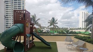 A playground with a slide and climbing structure, surrounded by outdoor seating and palm trees. - TOP/4BR/Million Dlls Sea Views/4 Balcony/Padel/Gym (Cancún)