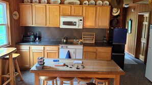 Een houten keuken met een magnetron, fornuis en koelkast. Er staat een tafel met krukken en diverse keukengerei erop.