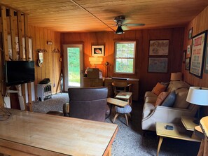 Un accogliente interno di una baita con soffitto in legno, una ventola a soffitto, un televisore, una scrivania con una sedia, un divano e quadri appesi alle pareti.