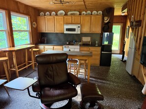 A compact kitchen with wooden cabinets, a microwave, a refrigerator, and a dining area with a chair and stools. - Spacious, waterfront privacy in Lake Leelanau , the heart of Leelanau County (Lake Leelanau)