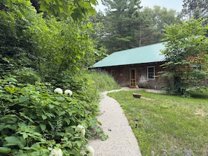 A rustic wooden cabin with a green roof, surrounded by dense greenery and a gravel path leading to the entrance. - Spacious, waterfront privacy in Lake Leelanau , the heart of Leelanau County (Lake Leelanau)