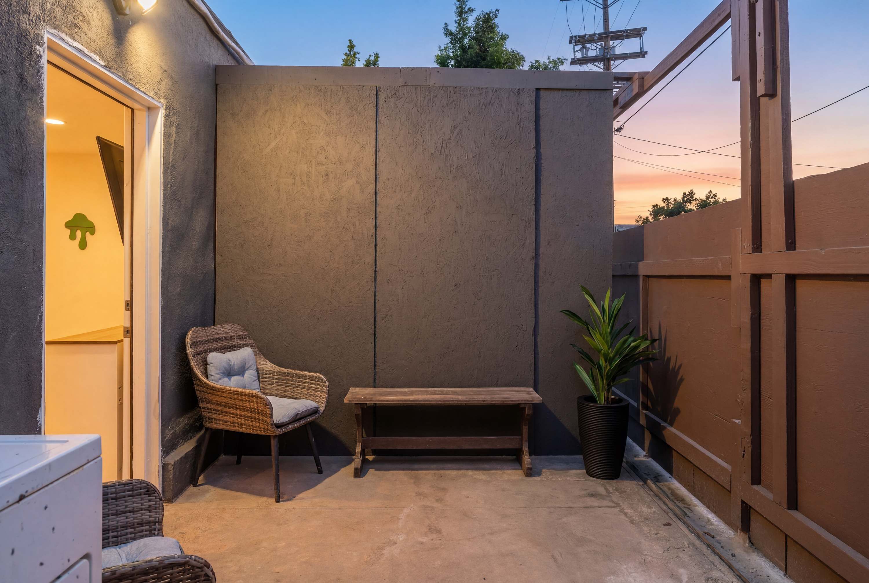 Un petit espace extérieur aménagé avec une chaise en osier et un banc, un pot de fleurs et un sol en béton.