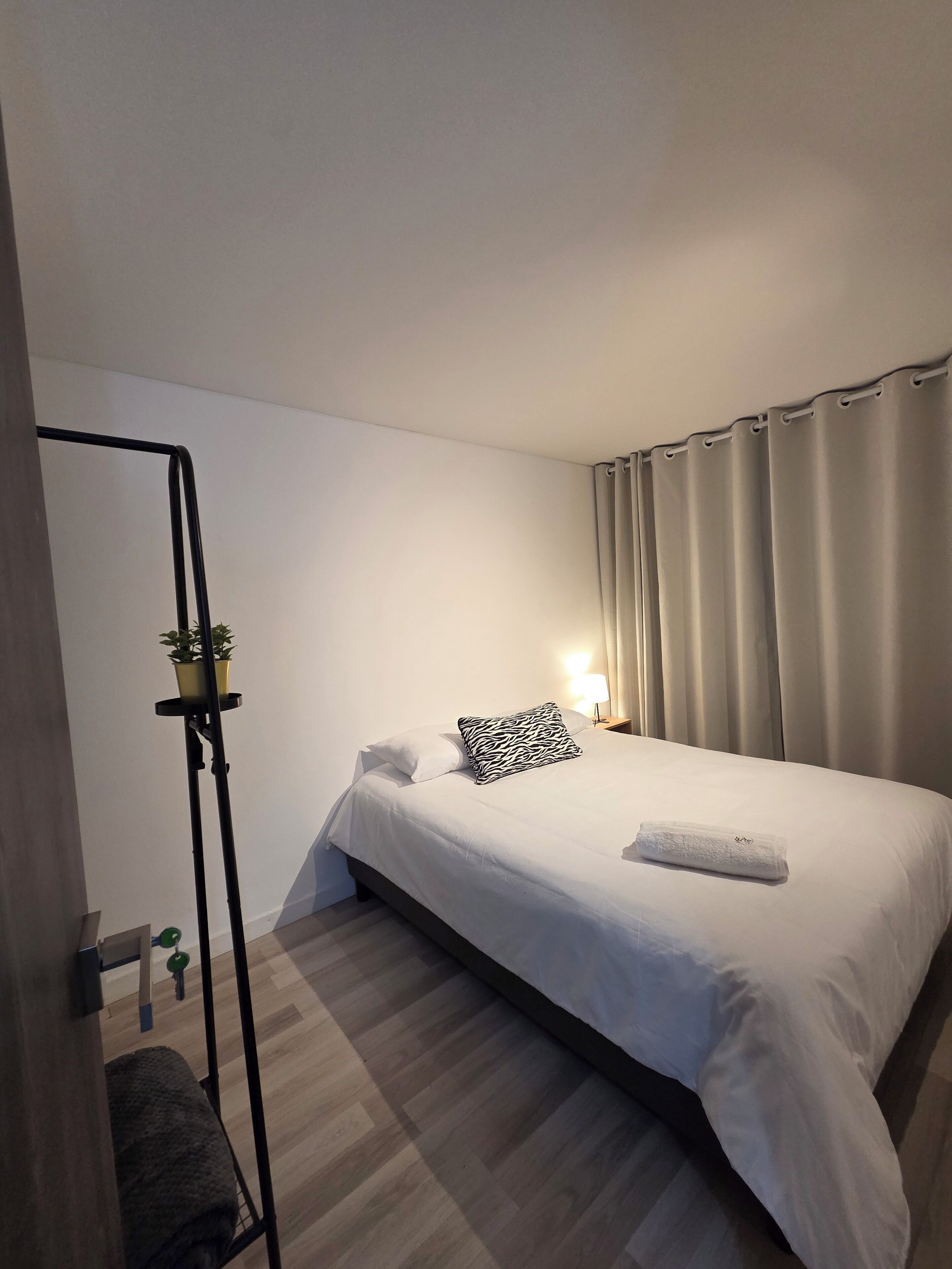 Hotellihuone, jossa on sänky, yöpöytä lampun kanssa, ruukkukasvi ja lattiavalaisin.