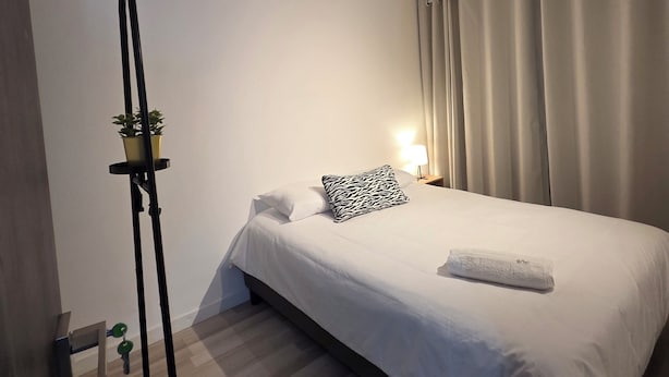 Une chambre d’hôtel avec un lit, une table de chevet avec une lampe, une plante en pot et un lampadaire.