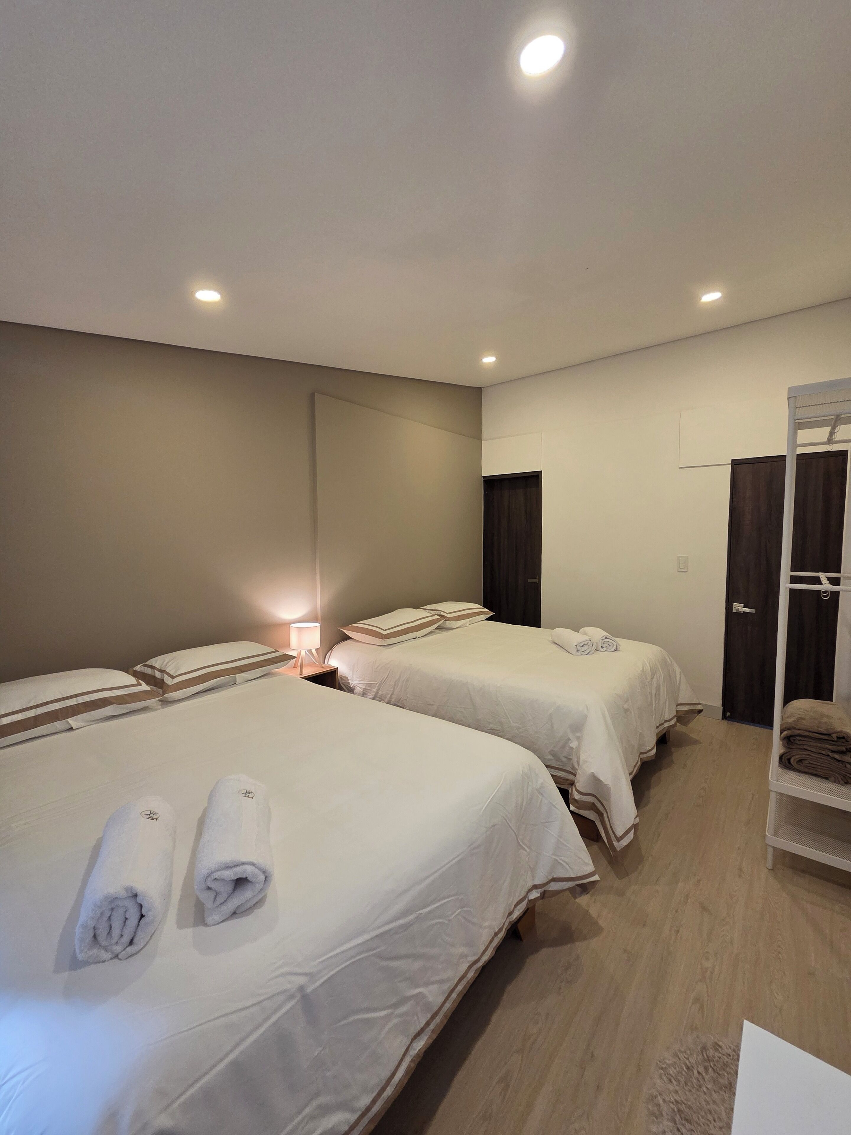 A hotel room with two beds, each with neatly folded towels, a bedside table with a lamp, and a wardrobe.