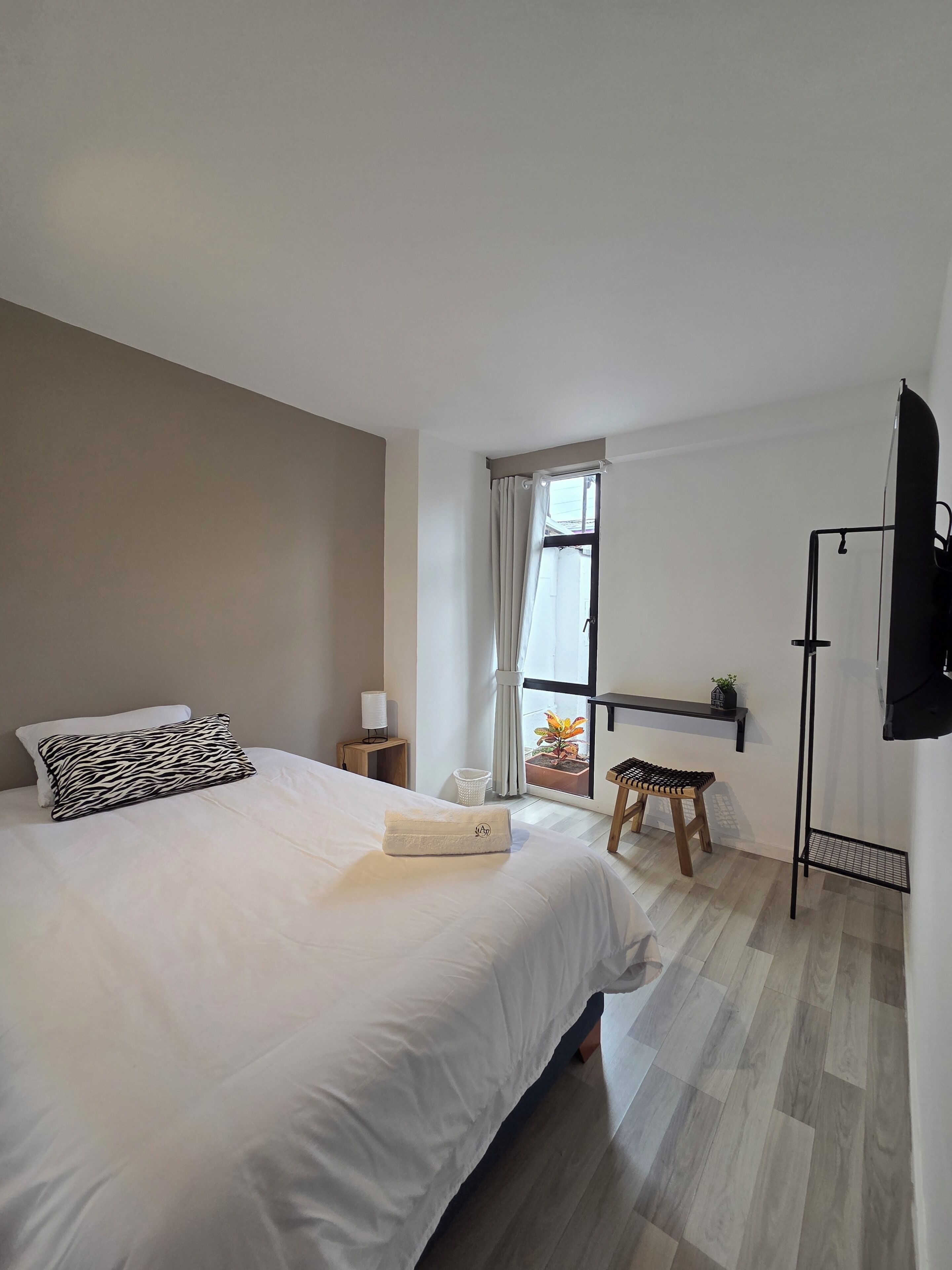 A bedroom with a bed, a nightstand, a window with curtains, a small stool, and a TV mounted on the wall.