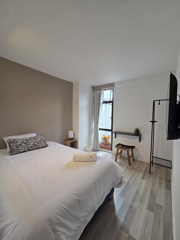 A bedroom with a bed, a nightstand, a window with curtains, a small stool, and a TV mounted on the wall.