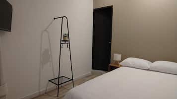Habitación de hotel con una cama, una mesita de noche, una lámpara, un estante de metal negro con plantas y una puerta.