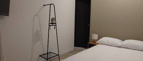 Habitación de hotel con una cama, una mesita de noche, una lámpara, un estante de metal negro con plantas y una puerta.