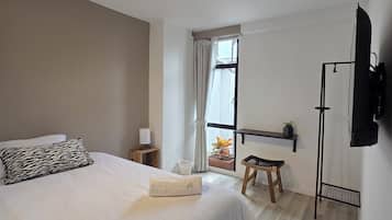 Un dormitorio con cama, mesita de noche con lámpara, ventana con cortinas, un taburete pequeño y televisor montado.