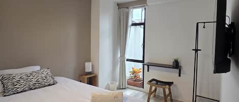 A bedroom with a bed, a bedside table with a lamp, a window with curtains, a small stool, and a mounted TV.