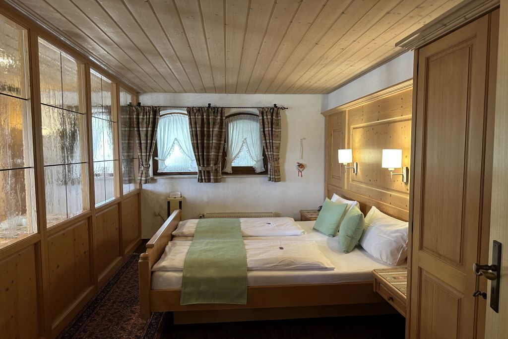 A room with a large bed, a wooden wardrobe, a window with curtains, and a wall-mounted lamp.