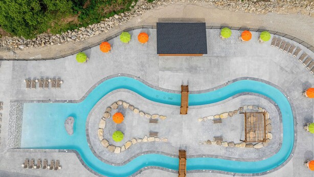 Luftaufnahme eines Schwimmbeckenbereichs mit mehreren Bahnen, Holzterrasse und umgebender Bepflanzung.