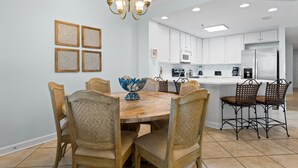 A dining area with a wooden table and chairs, a chandelier, and a kitchen in the background. - Beautiful Beach Front Condo With Breathtaking Views (Orange Beach)