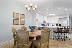 A dining area with a wooden table and chairs, a chandelier, and a kitchen in the background. - Beautiful Beach Front Condo With Breathtaking Views (Orange Beach)