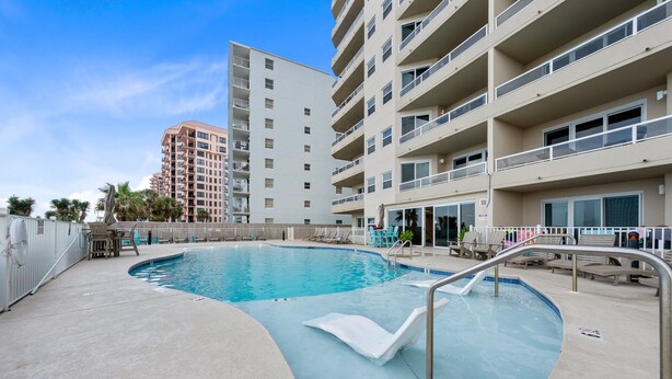 A swimming pool with a diving board, surrounded by lounge chairs and a building with multiple balconies. - Beautiful Beach Front Condo With Breathtaking Views (Orange Beach)