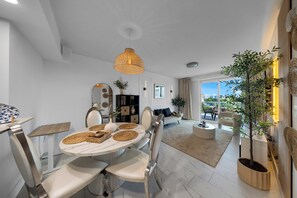 Ein modernes Wohnzimmer mit Essbereich, ausgestattet mit einem runden Tisch mit gewebten Platzsets, weißen Stühlen, einer Pendelleuchte und Blick ins Freie durch große Fenster.