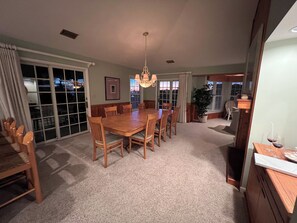 A dining room with a large wooden table surrounded by chairs, a chandelier, and a view through a sliding glass door. - Oceanside resort, hot tub, pool, walk to beach, tennis, amazing sunset views (Kitty Hawk)
