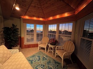 A cozy room with a wooden ceiling, large windows with blinds, a potted plant, a wicker chair, a small table with a vase, and a patterned rug. - Oceanside resort, hot tub, pool, walk to beach, tennis, amazing sunset views (Kitty Hawk)