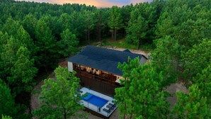 A two-story cabin surrounded by dense forest, with a visible deck and a swimming pool. - Record Time | Pool, Hot Tub, Game Room, Sleeps 15 (Broken Bow)