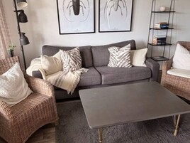 Una sala de estar con un sofá gris, dos sillones, una mesa de centro, una lámpara de pie y una estantería.