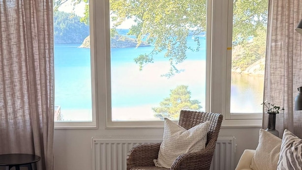 A room with a view of a lake, a wicker chair, a white sofa, and a black lamp. - 5 Star Holiday Home in Uddevalla (Uddevalla)