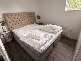 Un dormitorio con cabecera capitonada, una cama con ropa blanca, una mesita de noche con lámpara y una ventana con vistas a zonas verdes.