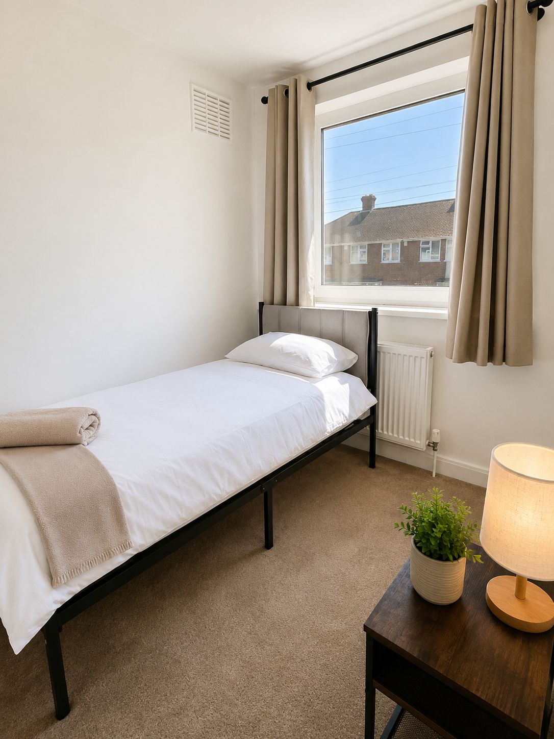 A single bed with a headboard, a window with curtains, a radiator, a table with a lamp and a plant, and a carpeted floor.