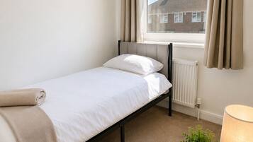 A single bed with a headboard, a window with curtains, a radiator, a table with a lamp and a plant, and a carpeted floor.