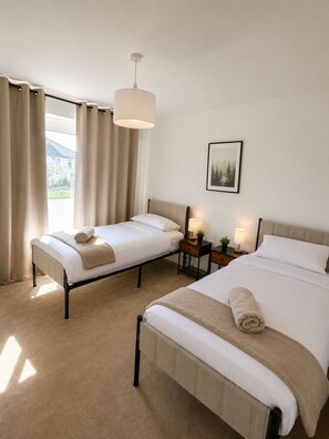 A hotel room with two beds, a large window with curtains, a hanging light fixture, a bedside table with a lamp, and a framed picture on the wall. - 4 BR Home - Parking, Garden, 9min to Universal Site (Bedford)
