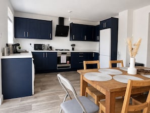 A modern kitchen with dark blue cabinets, a central island, and a dining area with a wooden table and chairs. - 4 BR Home - Parking, Garden, 9min to Universal Site (Bedford)