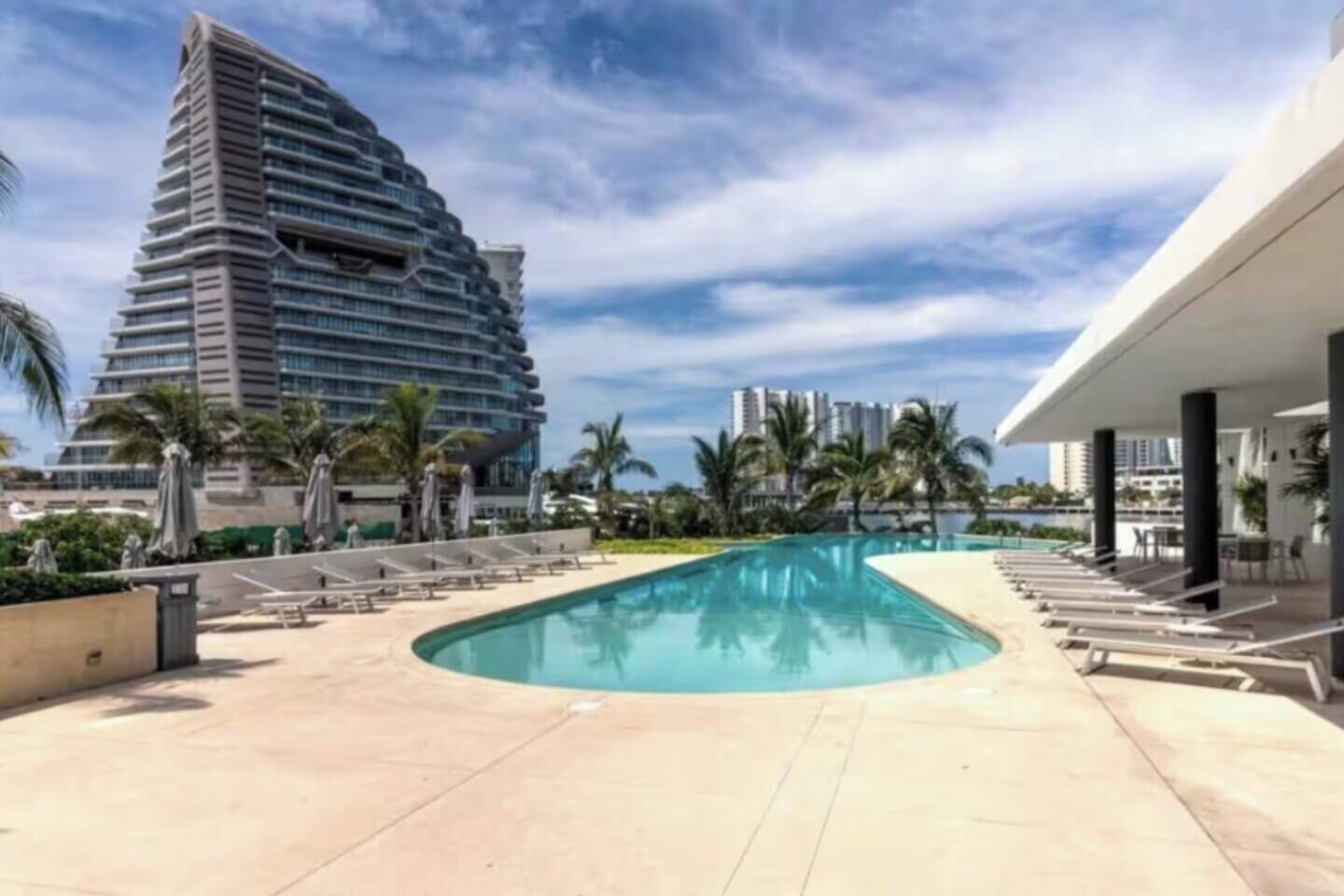A swimming pool with lounge chairs and umbrellas, surrounded by palm trees and modern buildings.