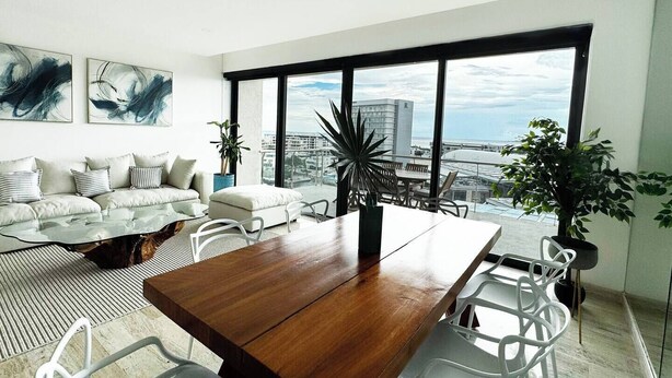 A modern living room with a large dining table, a sofa set, and a view of the city through large windows. - TOP/2BR/Home Away From Home/Ocean views/Balcony/HZ (Cancún)