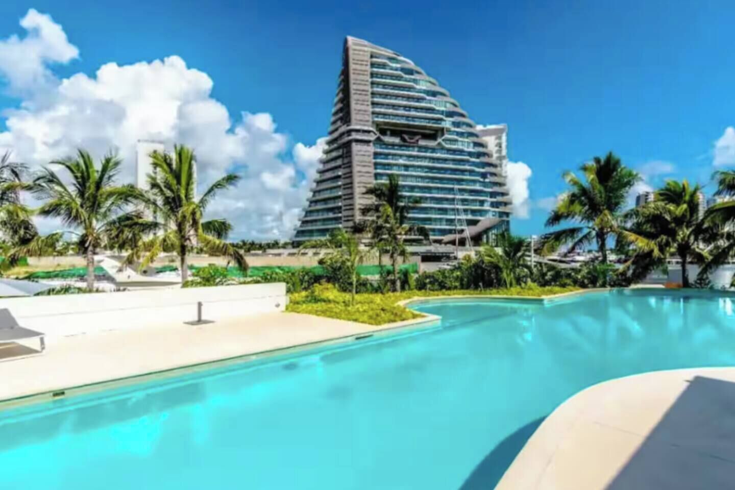 A modern hotel with a pool, palm trees, and a clear blue sky.