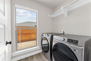 A laundry room with two washing machines, a window with a view of a wooden fence and a building, and a white door. - Unforgettable 4BD Oasis in West Texas (Midland)