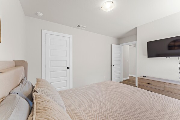 A bedroom with a bed, a TV mounted on the wall, and a door leading to another room. - Unforgettable 4BD Oasis in West Texas (Midland)