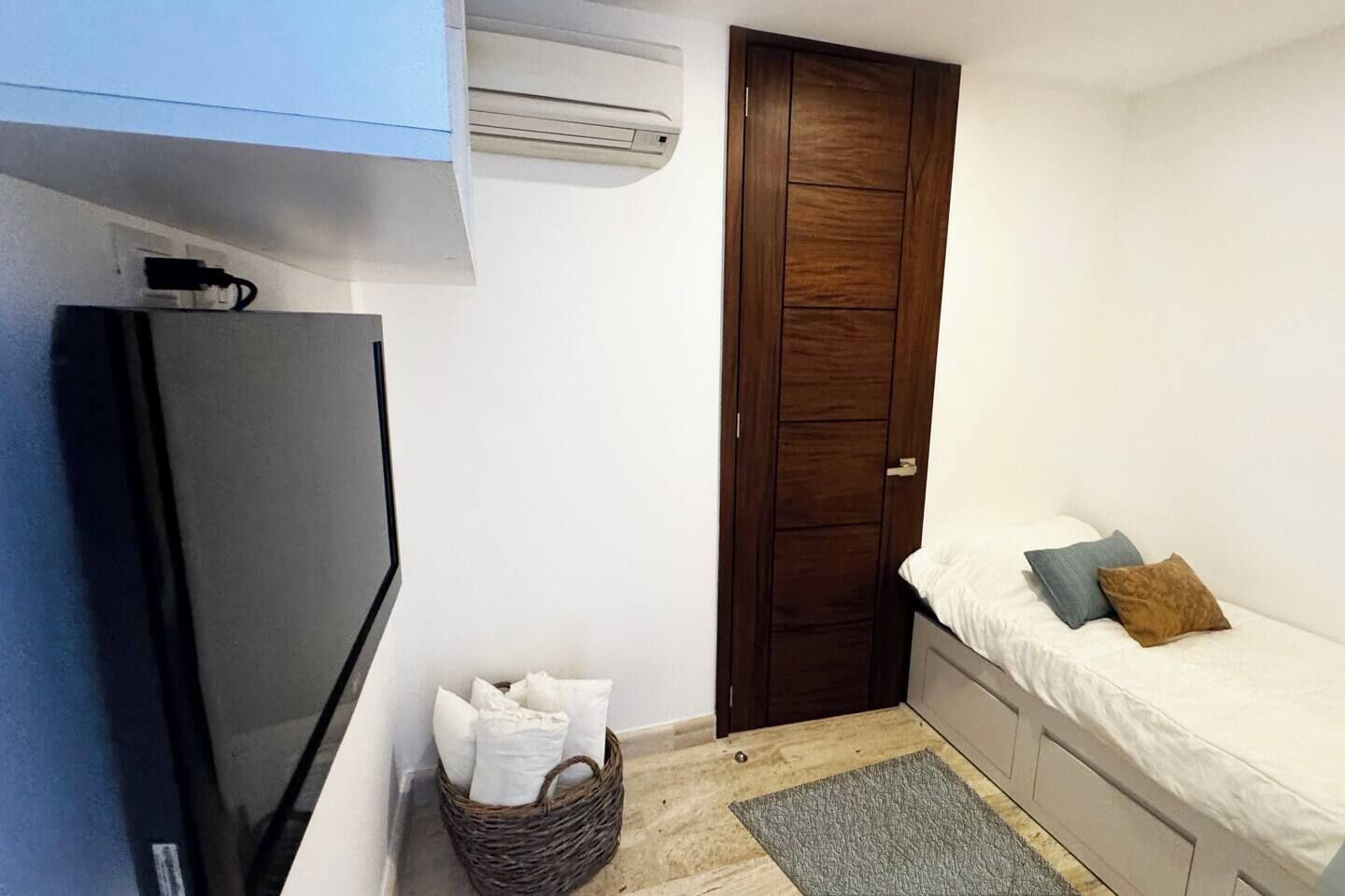 Ein kleiner, gemütlicher Raum mit Bett, Fernseher, Kleiderschrank und einem Kissenkorb.