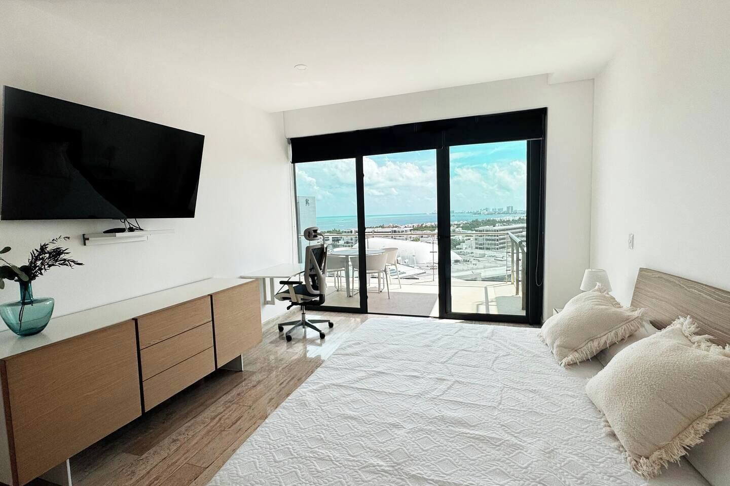 Une chambre moderne avec un grand lit, un téléviseur fixé au mur, un bureau avec une chaise et un balcon offrant une vue sur la ville et l’océan.